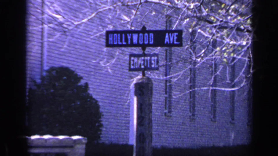 1968 Florida 8mm frame with Hollywood Ave and Em/ett St signs, a signpost, brick exterior, shrubbery, and bare tree limbs.