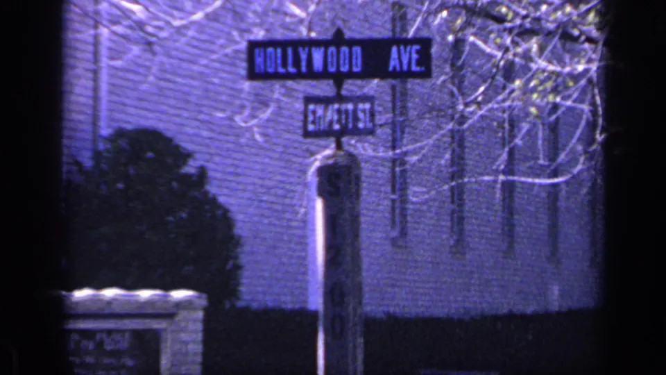 1968 Florida 8mm frame of Hollywood Ave and Em/ett St signs, with a brick building, dark shrub, low wall, and bare branches nearby.
