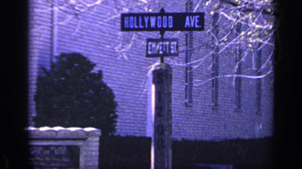 1968 Florida 8mm frame showing the Hollywood Ave street sign and cross-street sign against a brick wall, shrub, and pale bare branches.