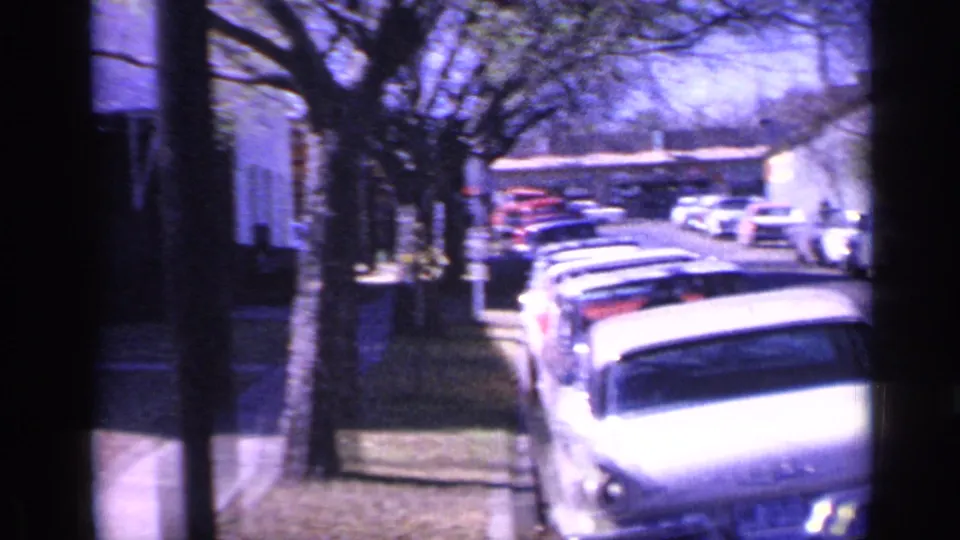 1968 Florida home-movie frame showing parked cars along a tree-lined residential street with small distant pedestrians on the sidewalk.