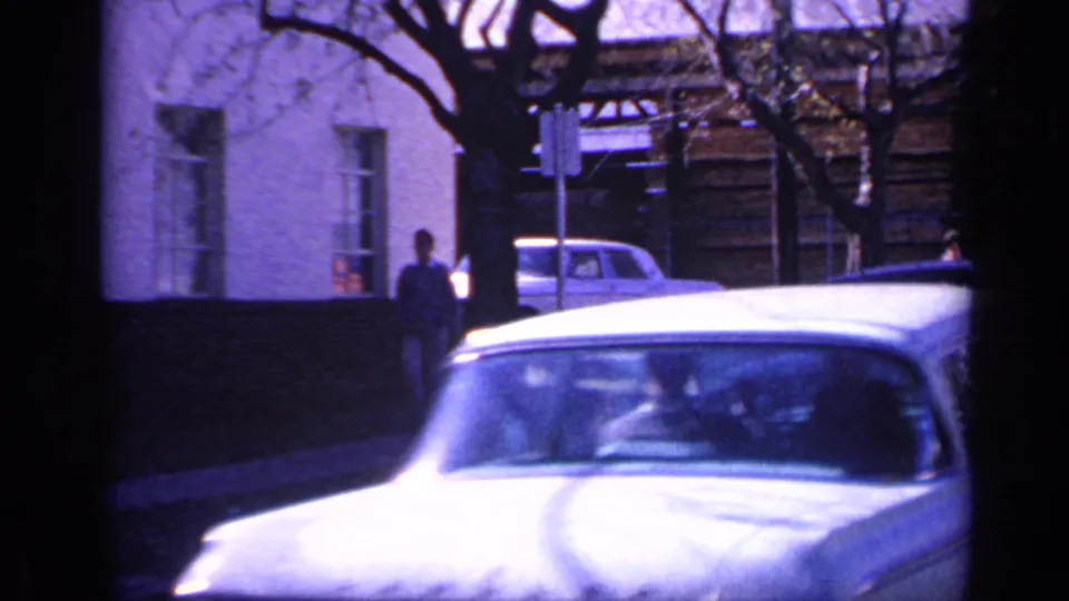 1968 Florida home-movie frame showing a light-colored car moving past a white building, with a pedestrian near a tree and street sign.