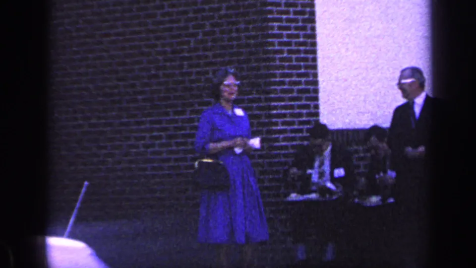 In 1968 Florida home-movie footage, a woman in a blue dress and glasses stands near a red brick wall with a purse in hand.