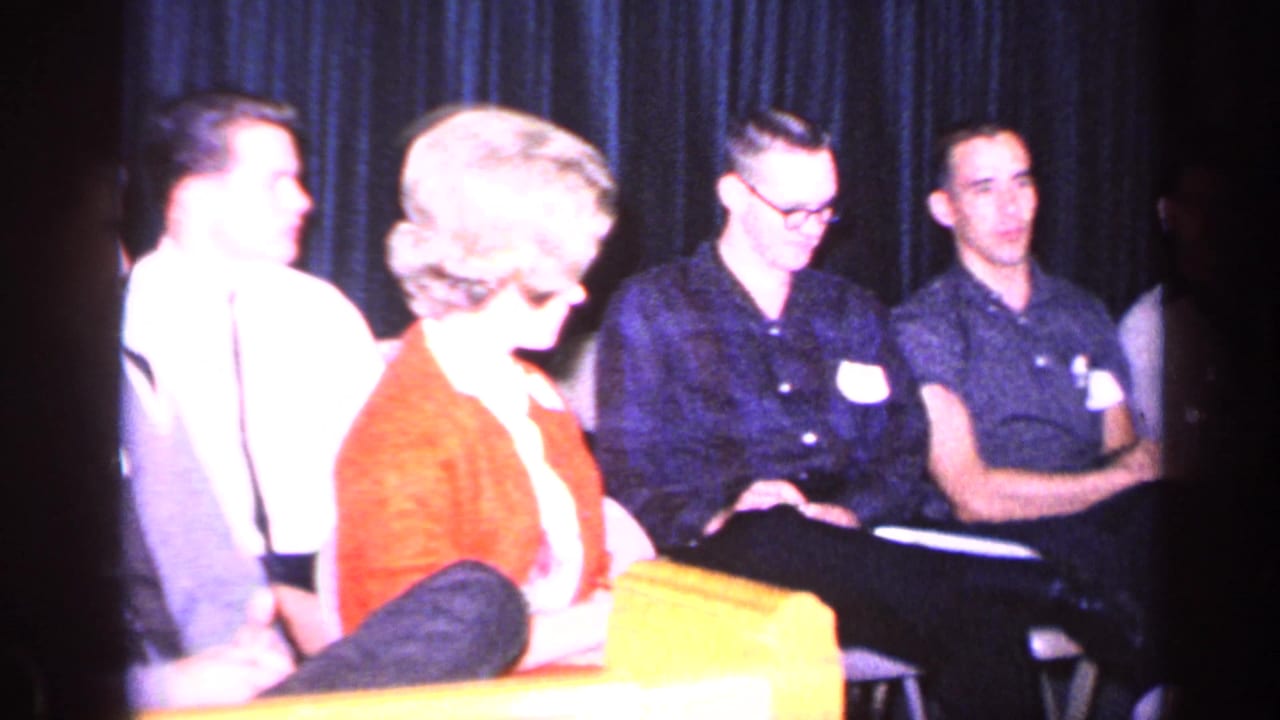 Several young adults sit in an auditorium setting, including a woman in a red cardigan and men in collared shirts.