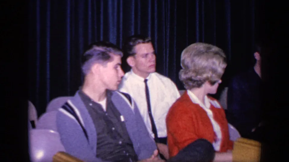 A man in a patterned shirt, a man in a white shirt and tie, and a woman in a red cardigan sit together indoors.