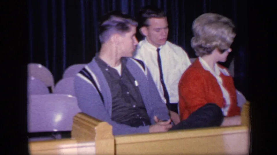 The seated group turns toward something off-camera, with pale seats, a yellow rail, and a dark curtain visible behind them.