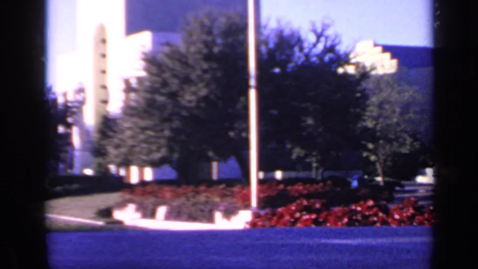 1968 Florida home-movie frame with sunlit white architectural forms, trees, flagpoles, red planting beds, and a reflective pool edge.