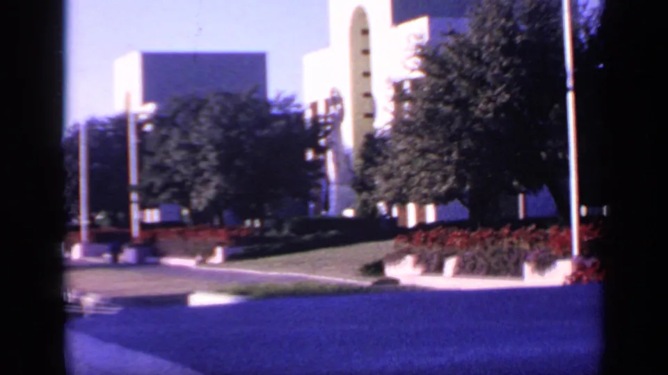 1968 Florida 8mm frame looking across a landscaped walkway, flagpoles, trees, red flower beds, grass, and white modernist buildings.