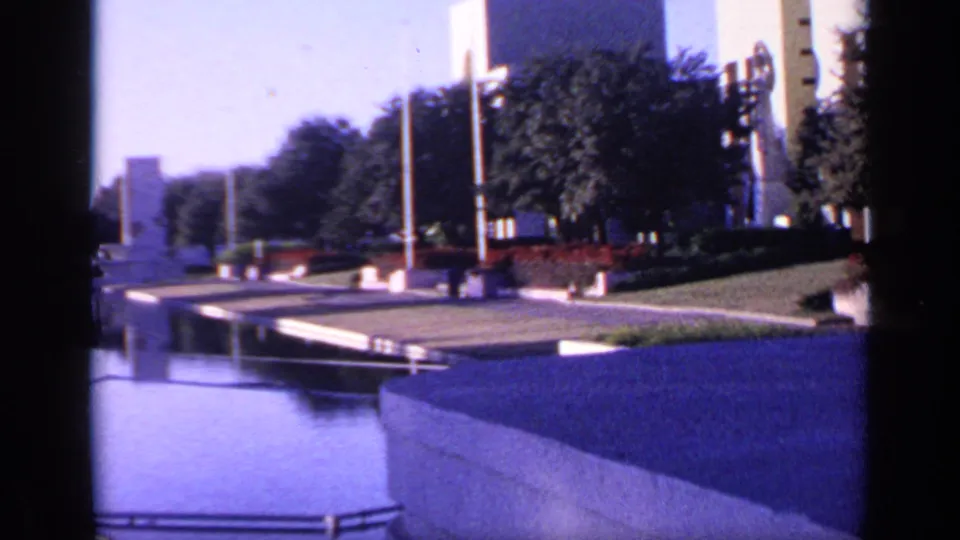 1968 Florida home-movie frame showing a reflecting pool and low wall in front of lawn, red flowers, flagpoles, trees, and white buildings.