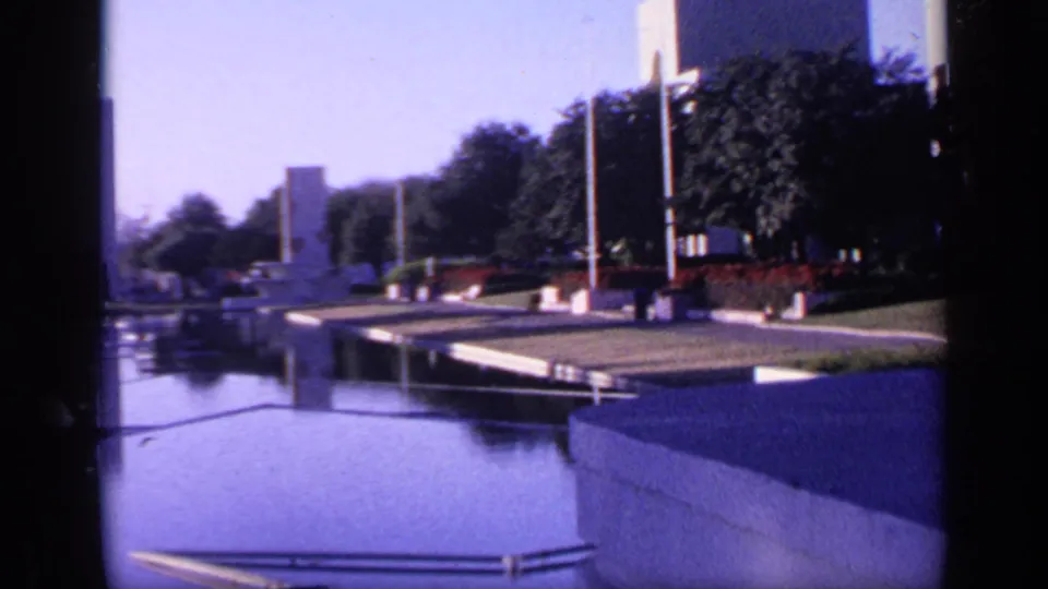 1968 Florida 8mm frame showing a calm reflective pool bordered by walkways, trees, railings, and vertical memorial-like monuments.