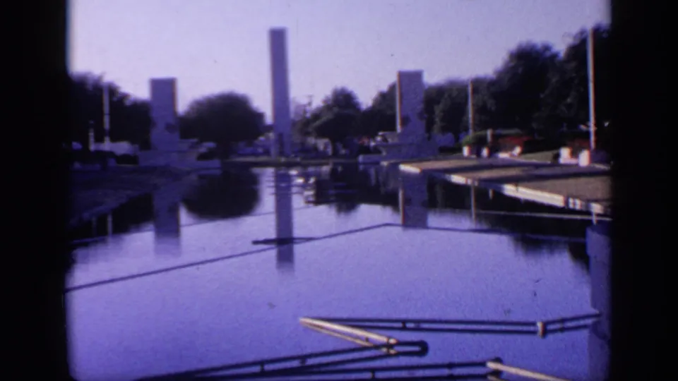 1968 Florida home-movie frame looking down a reflective pool or canal lined with trees, walkways, railings, and tall rectangular monuments.