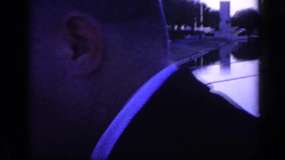 Close 1968 Florida home-movie frame of an adult man in profile, with reflective water, railings, trees, and monuments behind him.