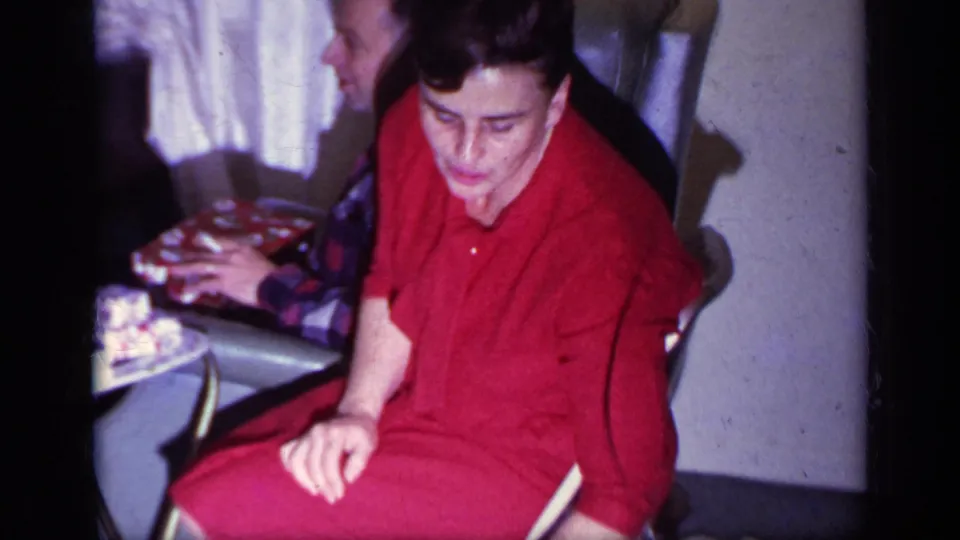 1969 California home-movie frame showing a woman in red seated indoors while a man in plaid sits partly behind her.