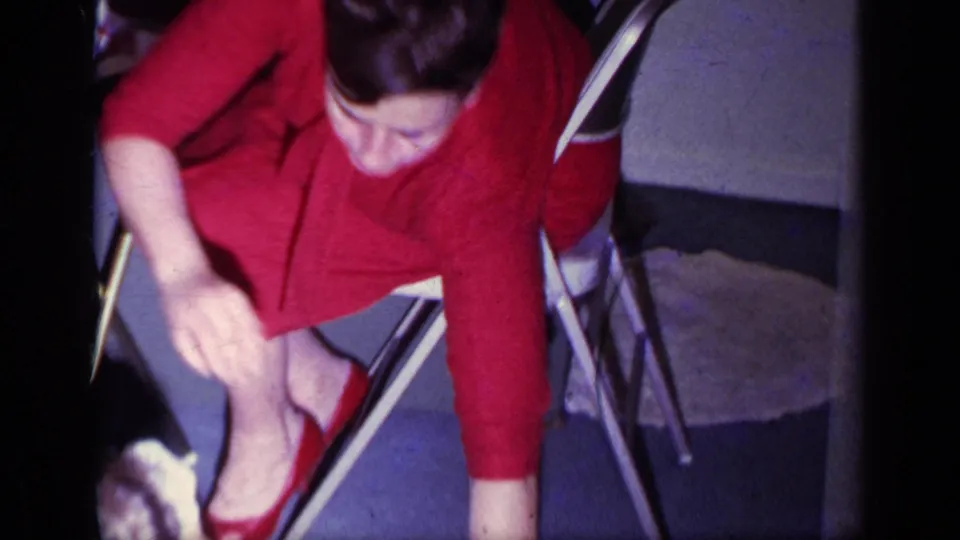 1969 California 8mm frame of a woman in a red outfit leaning forward from a folding chair inside a home.