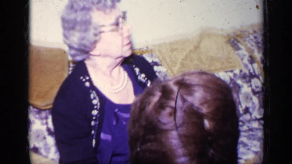 1969 California home-movie frame of an older woman with glasses, pearls, and dark cardigan seated on a patterned couch.