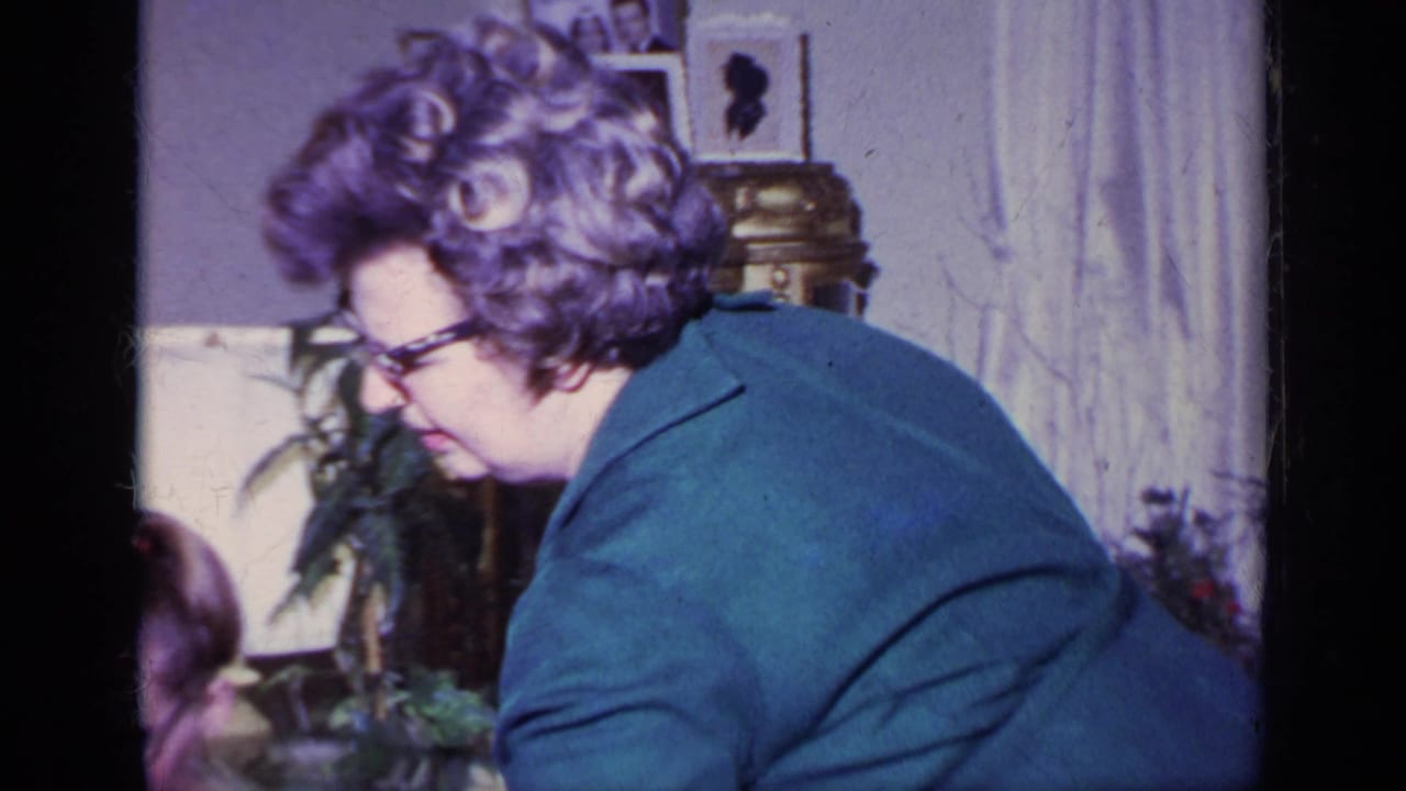 Gray-haired woman in glasses leans across a living room near a seated person.