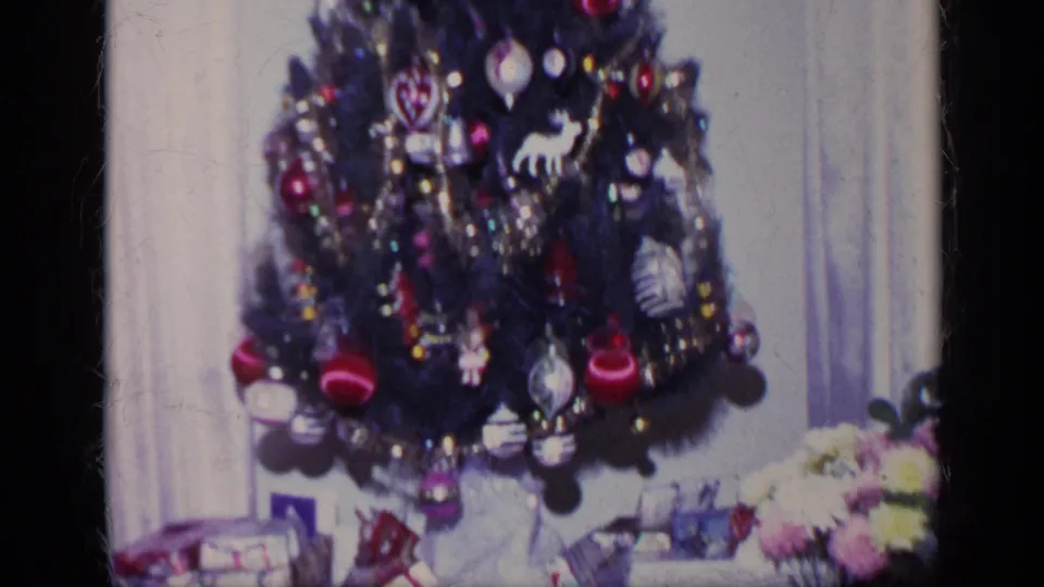 A decorated Christmas tree fills the frame, with red ornaments, tinsel, wrapped gifts, curtains, and a floral arrangement visible nearby.