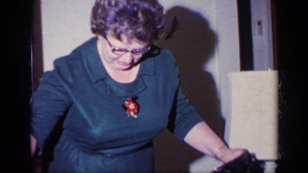 Woman in a blue dress and glasses looks down beside a table lamp.