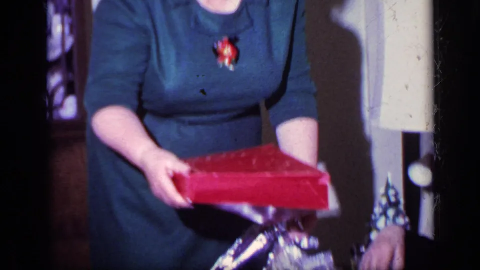 A woman in a blue dress holds a red gift box above shiny wrapping inside a California home setting from 1969.