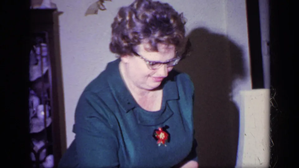 A woman in glasses and a blue dress looks down in a warmly lit California home interior, with a lamp and cabinet behind her.