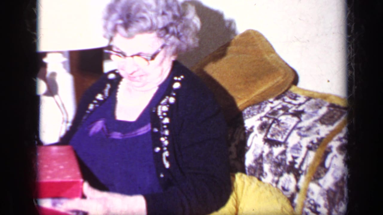 Older woman in glasses sits on a patterned couch in a California home-movie Christmas scene from 1969, holding a red gift box near a table lamp.