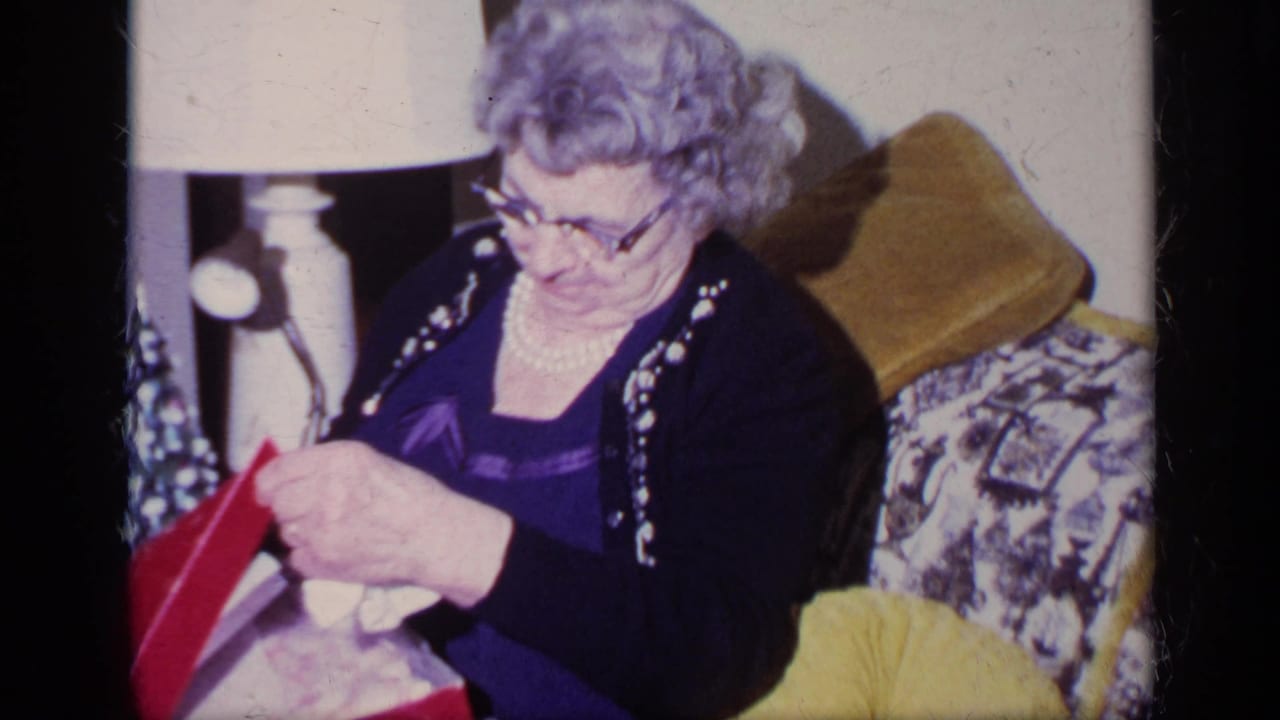 In this 1969 California home-movie frame, an older woman in a pearl necklace opens a red Christmas gift box on a patterned couch.