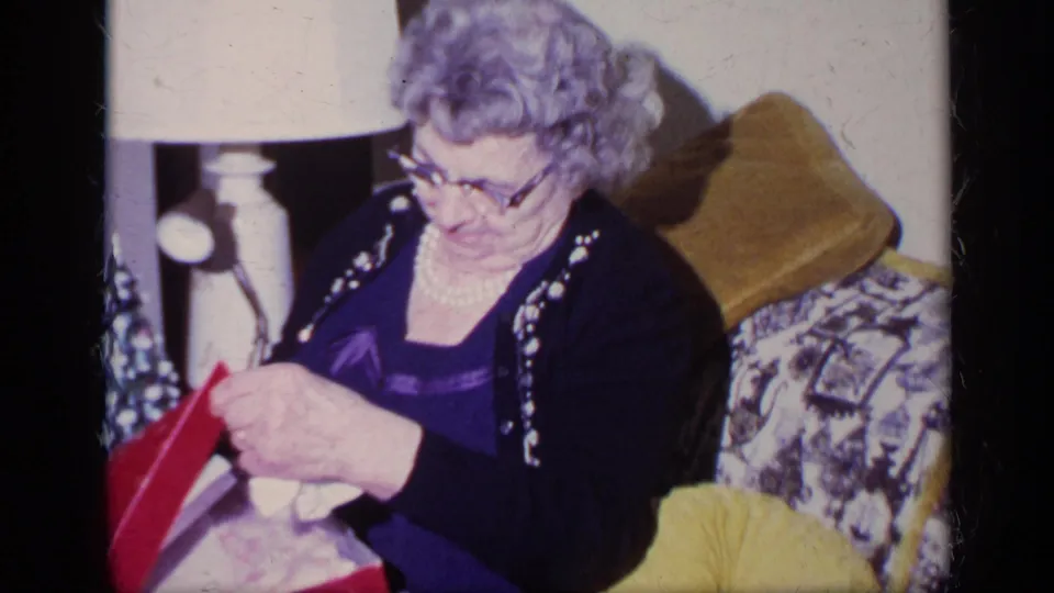 In this 1969 California home-movie frame, an older woman in a pearl necklace opens a red Christmas gift box on a patterned couch.