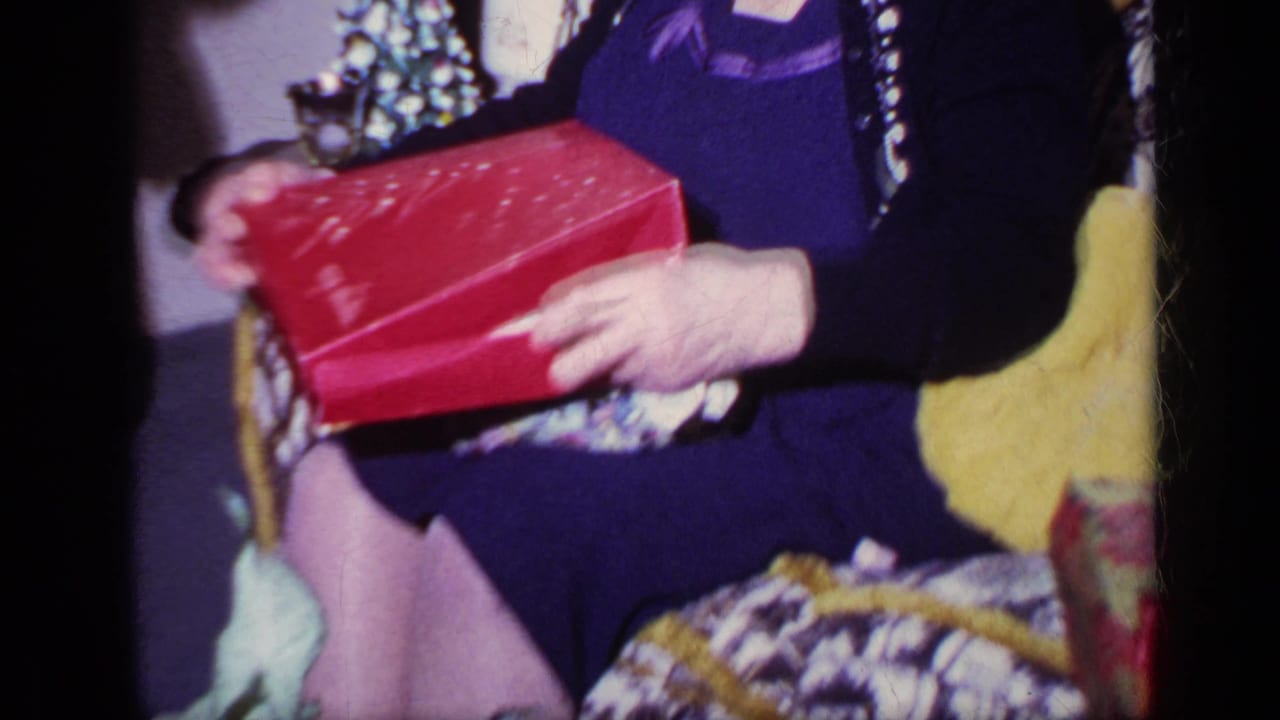 Close home-movie view of the seated woman's hands and lap as she handles a red Christmas gift box in a 1969 California living room.
