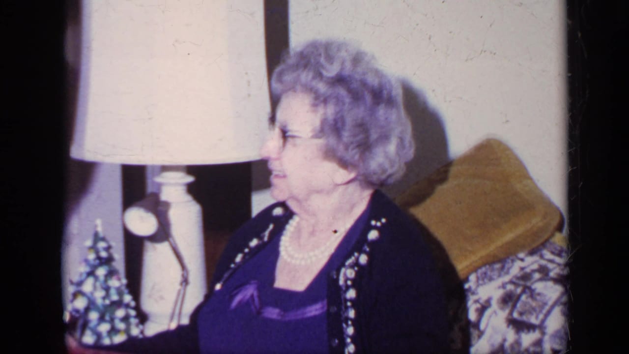 Older woman in glasses looks off to the side on a patterned couch, with a lamp and small Christmas tree visible in the 1969 California interior.