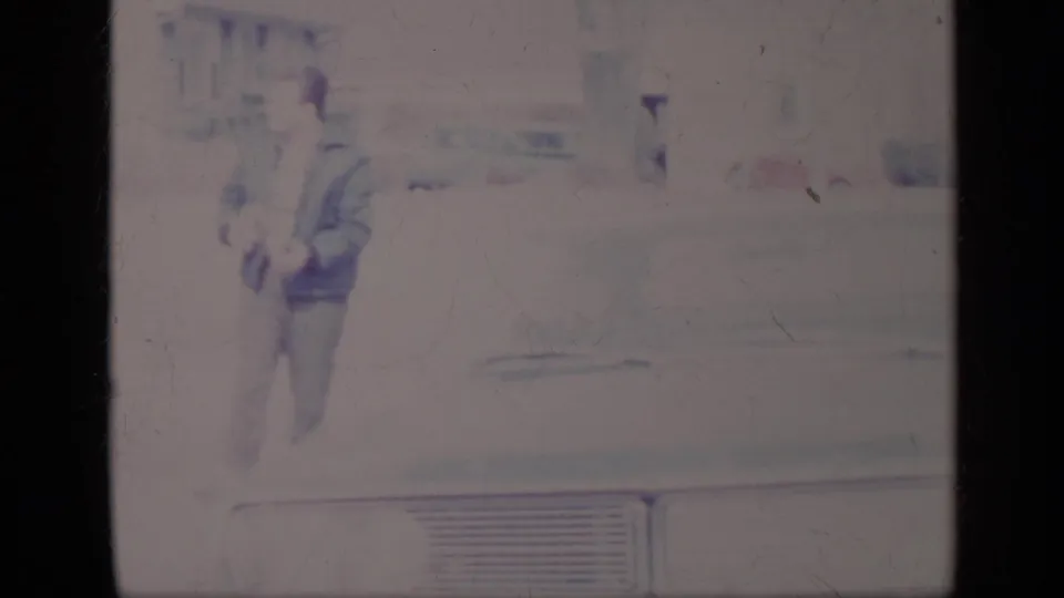 A man walks near a light-colored classic American car, with the faded 8mm frame emphasizing the vehicle's broad body and trim.