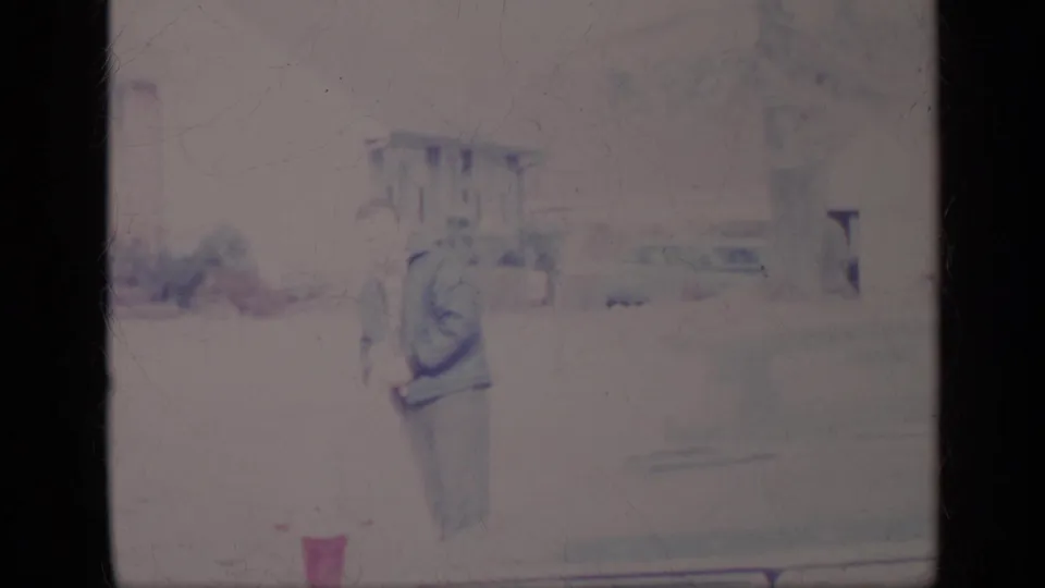 Faded 1969 California home-movie frame showing a man and smaller companion walking beside a parked car on a residential street.