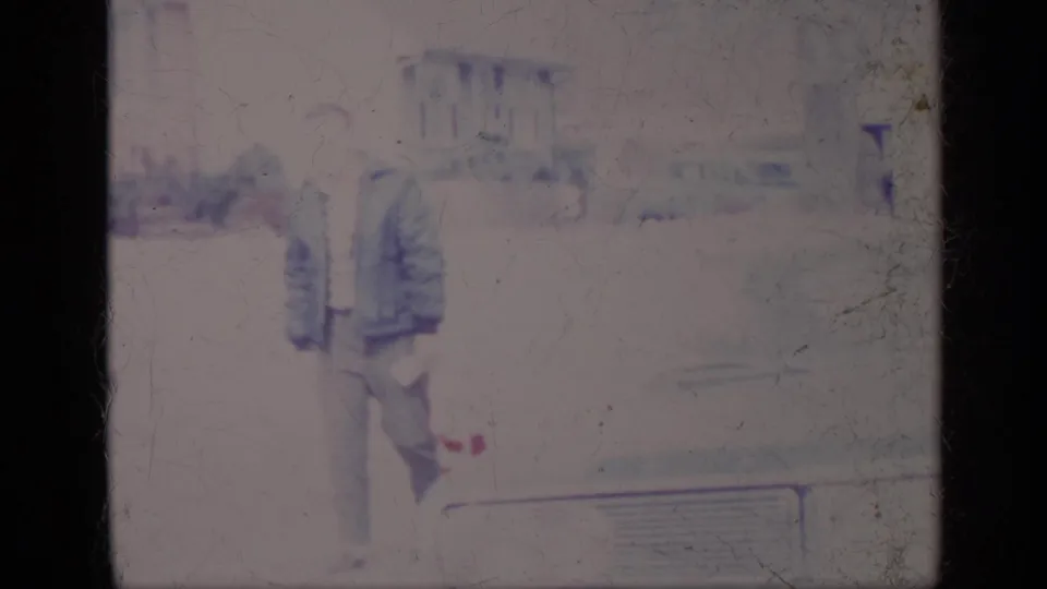 Soft 8mm frame from 1969 California showing a man in a jacket and smaller companion near the front of a parked car.