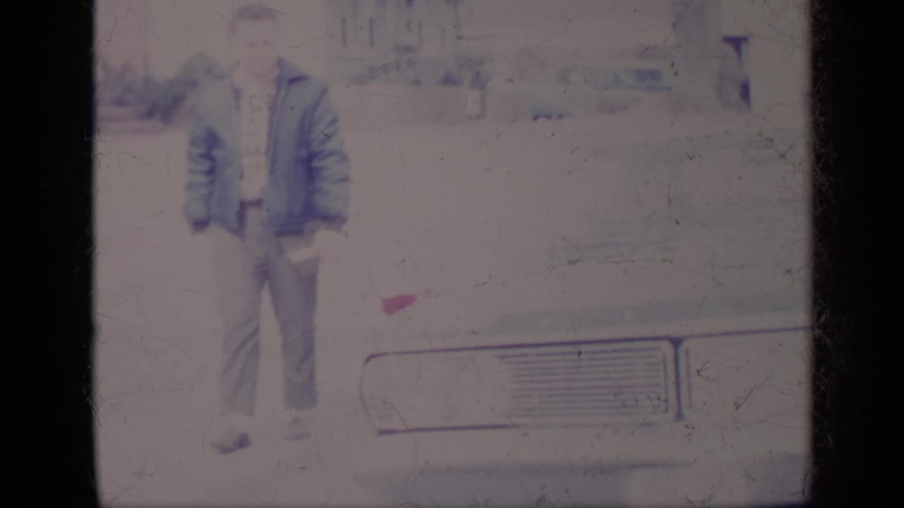 Man in a blue jacket standing near the front of a pale car in a driveway.