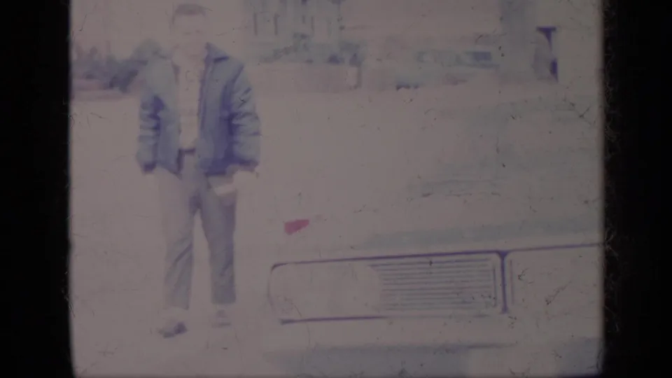 In a 1969 California home-movie frame, a man in a blue jacket stands beside the front of a pale car near a residential driveway.