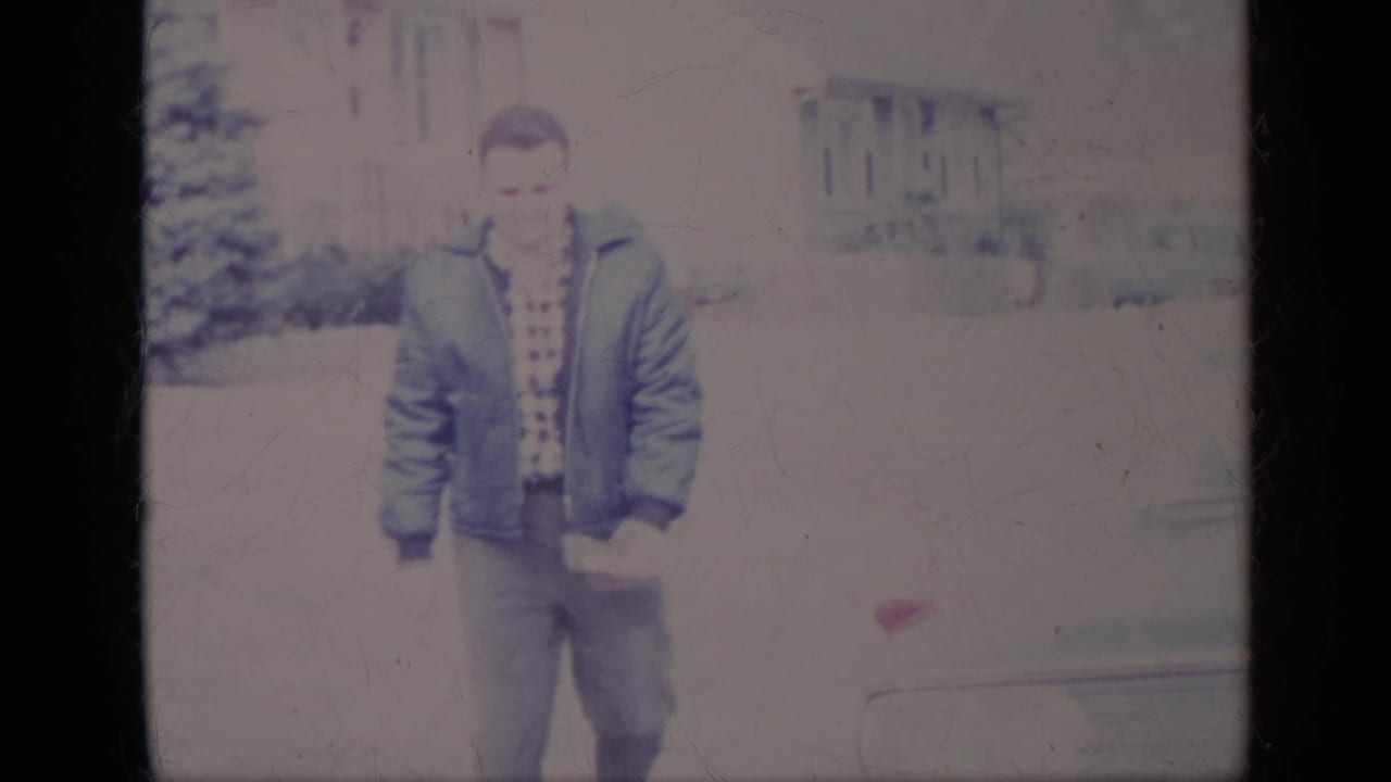 The man faces the camera in a blue jacket and patterned shirt, with part of a car and a house visible behind him in California.