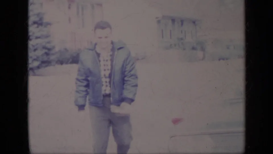 The man faces the camera in a blue jacket and patterned shirt, with part of a car and a house visible behind him in California.