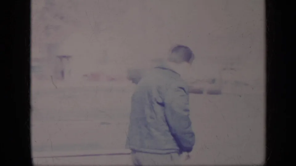 The man turns away from the camera near the pale car, with the bright residential background softened by 8mm exposure.