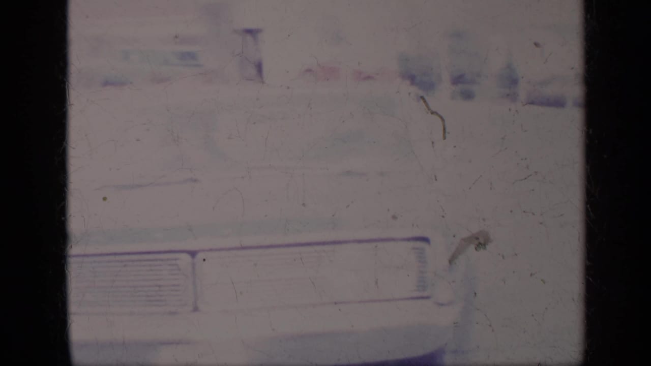 Faded 8mm frame showing the front corner of a white car with grille and dark film borders.
