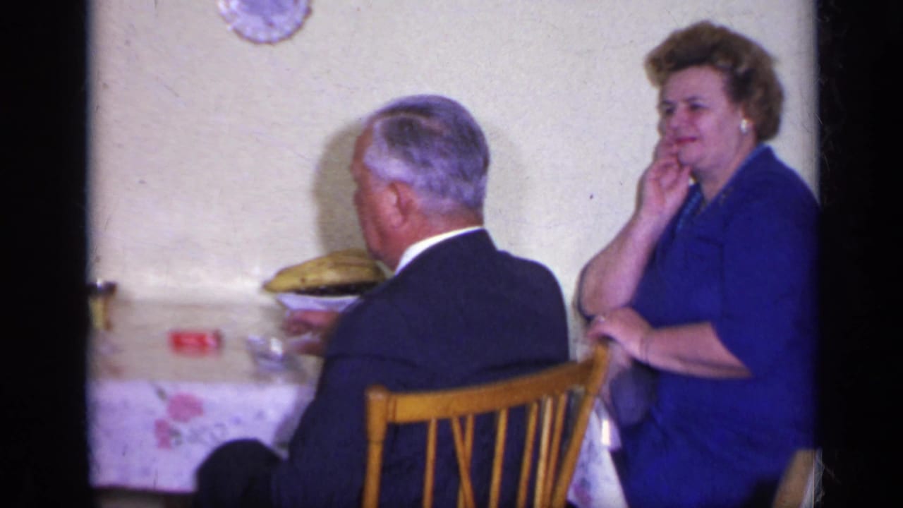 In a 1969 California home-movie frame, two adults sit at a dining table with chairs, dishes, and a wall clock visible.