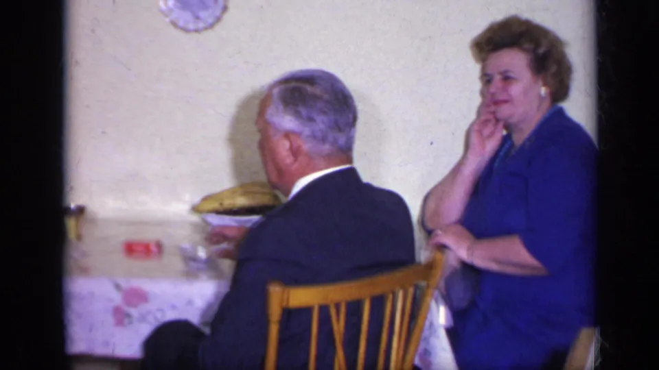 In a 1969 California home-movie frame, two adults sit at a dining table with chairs, dishes, and a wall clock visible.
