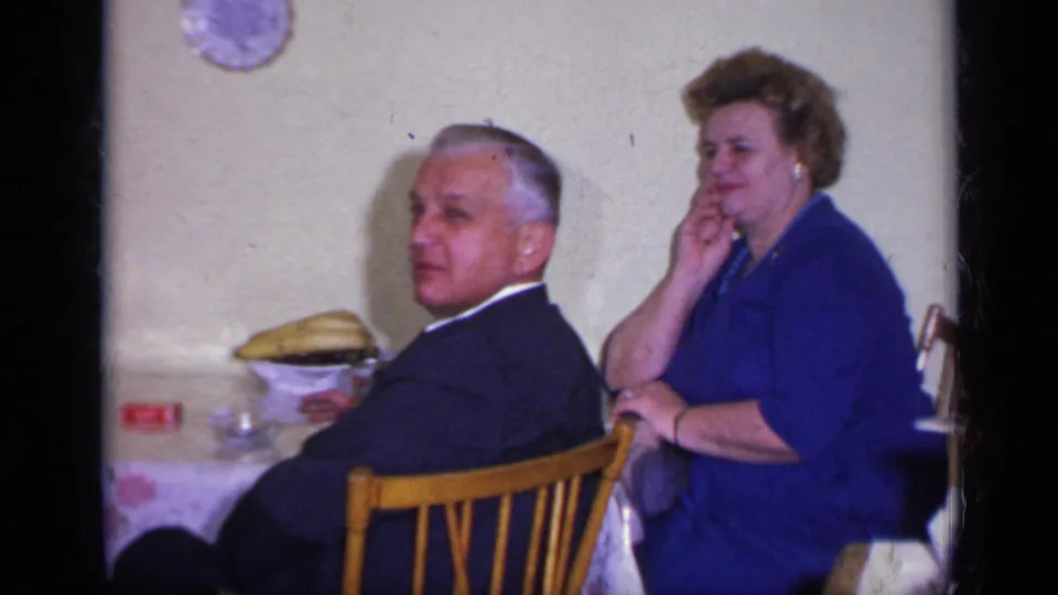 1969 California home-movie frame showing a suited man glancing toward the camera and a woman seated near a floral-covered table.