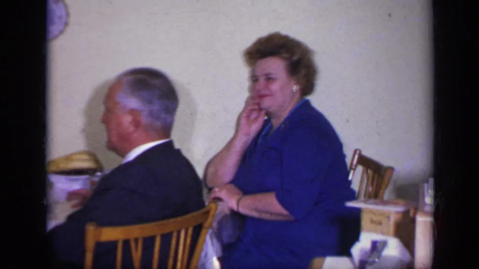 1969 California 8mm frame of a woman in a blue dress seated by a dining table while a suited man faces away.