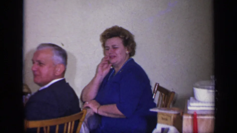 1969 California home-movie frame showing two adults seated near a floral-covered table in an indoor gathering.