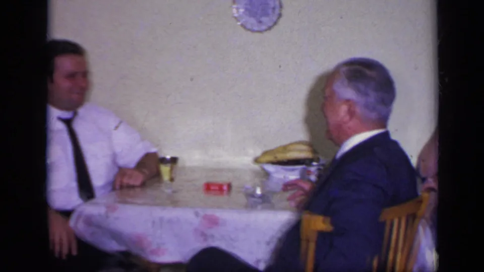 1969 California home-movie frame showing two men seated across a floral-covered table during a casual indoor gathering.