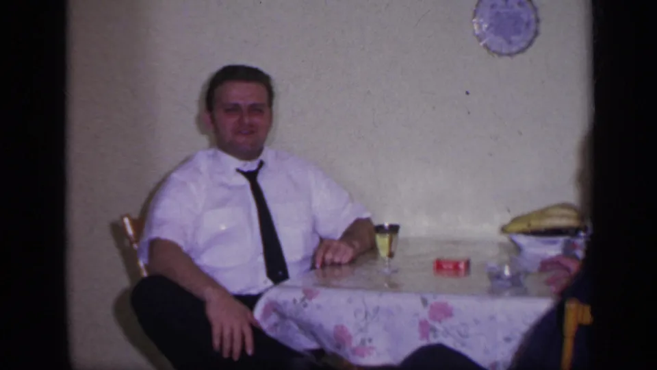 1969 California indoor frame showing a seated man in a white shirt and tie at a floral-covered table.