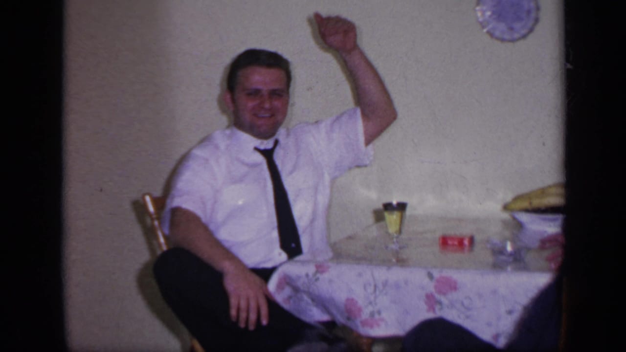 Man in a white shirt and black tie waves from a small table indoors.