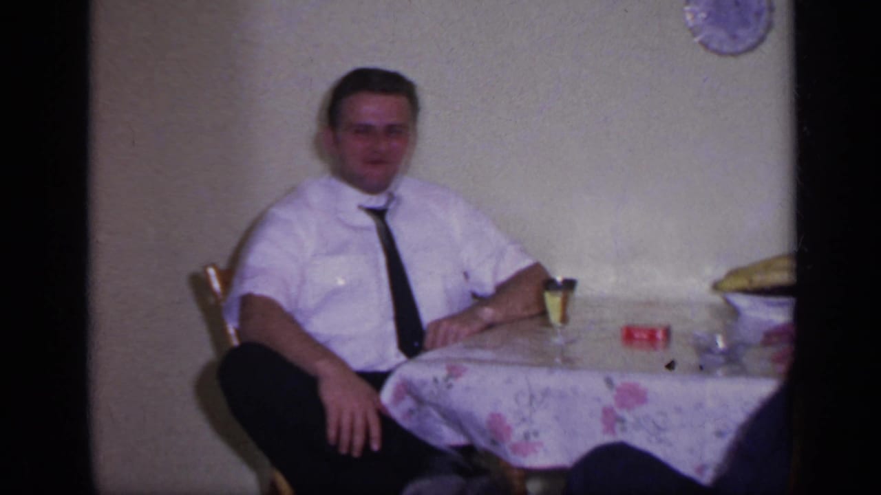 Man in a white shirt sits at a table with a glass and red item nearby.
