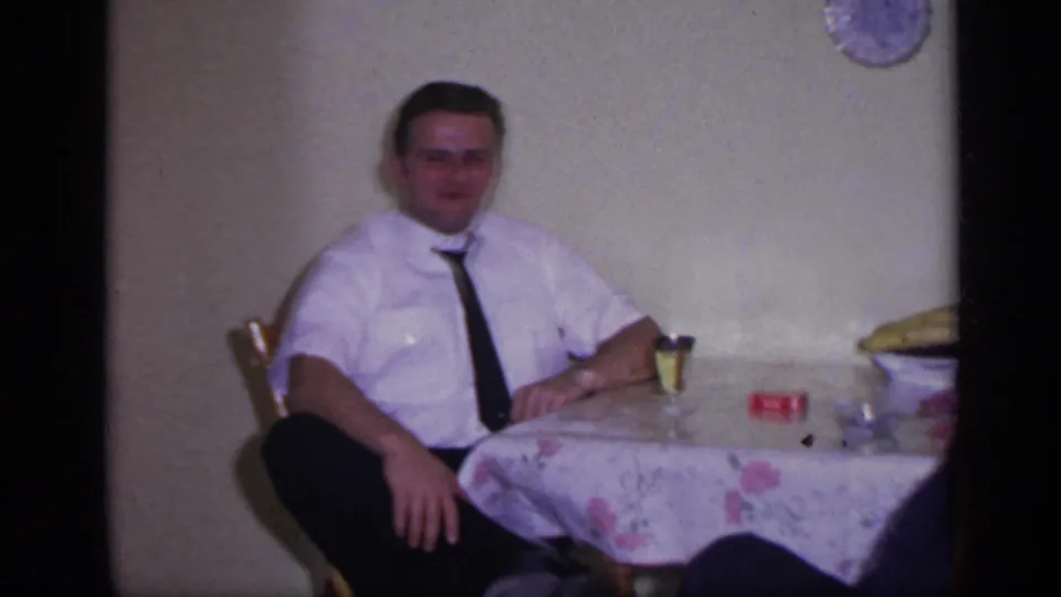 A seated man in a white shirt and black tie faces the camera at a floral-covered table in the 1969 California home movie.
