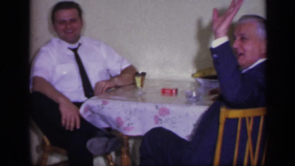 Two adult men sit around a small table, with the older man in a dark suit lifting a hand in this 1969 California home-movie frame.