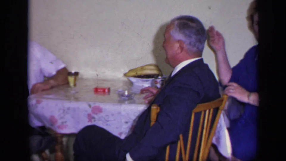 An older man in a dark suit sits side-on at the table while a woman in blue appears at right in the 1969 California home movie.