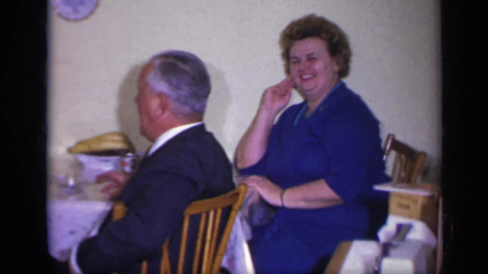 In the California home-movie scene, the woman in blue smiles toward the camera as the man turns away at the dining table.